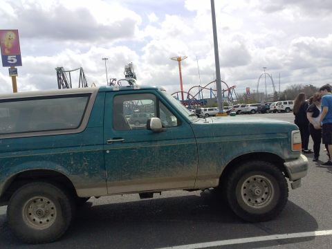 1996 Ford Bronco - A side profile of a 1996 Ford Bronco, seen parked in a lot with amusement park rides in the background.