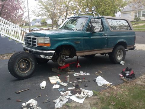 1996 Ford Bronco - A green 1996 Ford Bronco is parked on a driveway with its front wheel removed and supported by jack stands, surrounded by tools and parts.