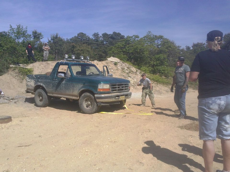 Green Ford Bronco with roof lights and yellow tow strap, parked on dirt with people around