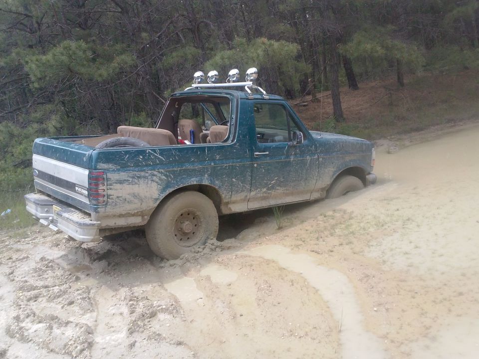 Mud-covered green Ford Bronco stuck in a dirt track, rear-left wheel submerged