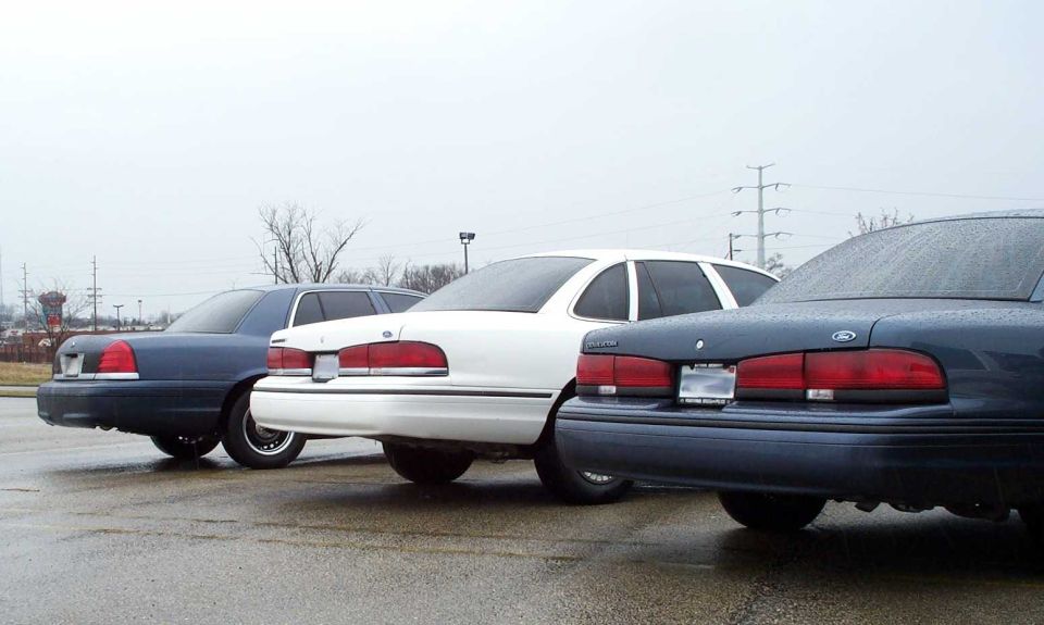 Three Ford Crown Victoria sedans parked rear-first, dark blue, white, and gray, on wet lot