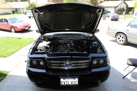 2003 Ford Crown Victoria - Front view of a black Ford sedan with its hood raised showing the engine bay and a California plate reading EVIL VIC, parked in a driveway.