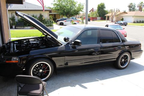 2003 Ford Crown Victoria - black full-size sedan with its hood open sits in a driveway outside an open garage, a folding chair to the left.