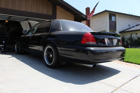 2003 Ford Crown Victoria - Rear three-quarter view of a black sedan with a trunk spoiler, dual exhaust tips and black wheels in a driveway.
