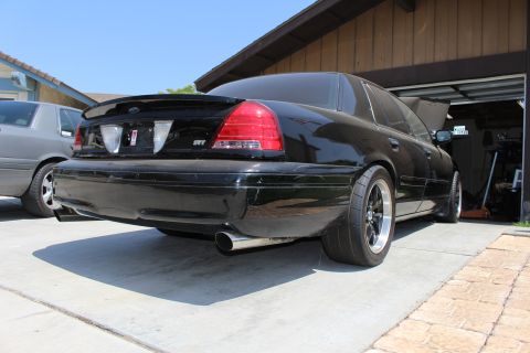 2003 Ford Crown Victoria - Rear three-quarter view of a black sedan with an SVT badge and chrome exhaust tip parked beside an open garage.