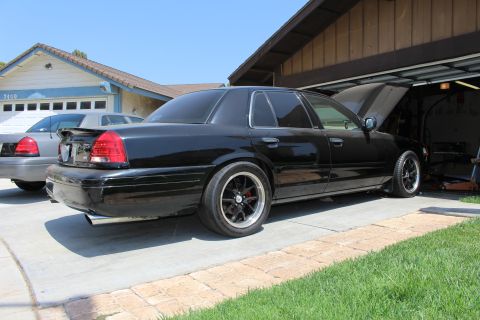 2003 Ford Crown Victoria - Rear three-quarter view of a lowered black sedan on black wheels in a driveway, a second sedan parked behind it.