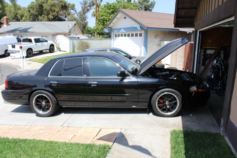 2003 Ford Crown Victoria - Full side profile of a lowered black four-door sedan with hood raised in a driveway, a white pickup parked across the street.