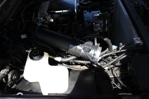 2003 Ford Crown Victoria - black intake pipe with a mass airflow sensor connecting to the supercharger, beside a coolant reservoir.