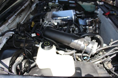 2003 Ford Crown Victoria - Engine bay dominated by a large black air intake tube routing to a supercharged motor, with a coolant reservoir at center.