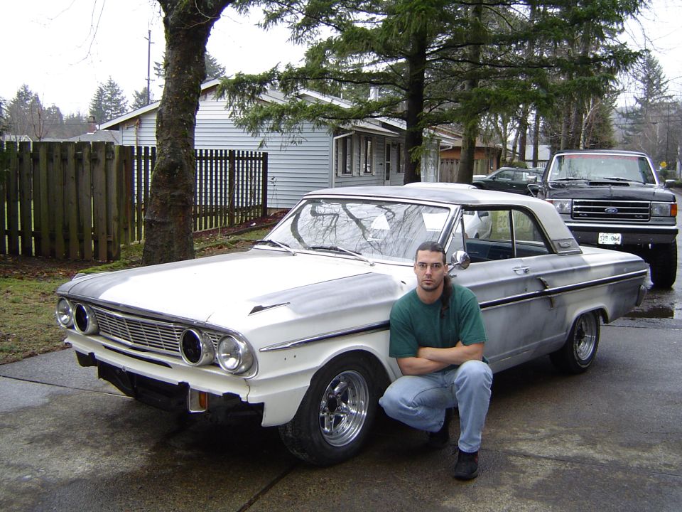 Man kneeling beside a white 1960s Ford Fairlane two-door with chrome wheels in a driveway