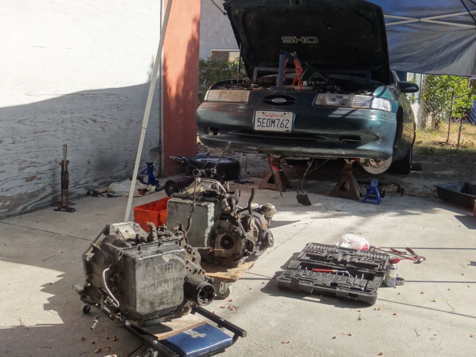 1993 Ford Taurus SHO on jack stands, hood open, transmission removed and on a dolly. Tools are on the ground
