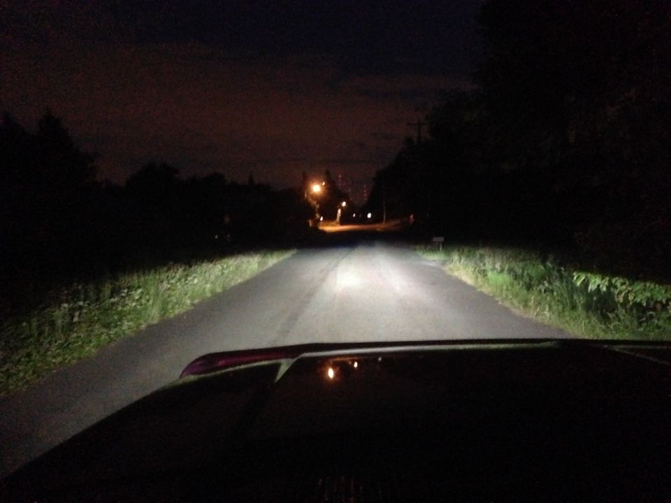 Vehicle hood visible in foreground, driving on a dark road at night with headlights on. Supermotors.net logo in lower right