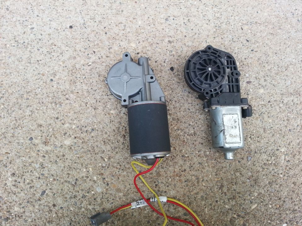 Two power window motors on concrete: aluminum-and-black motor with wires beside a black plastic and silver motor.