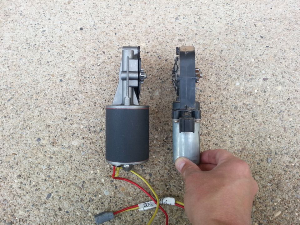 Two power window motors compared side by side on concrete, one with wiring, one held by hand