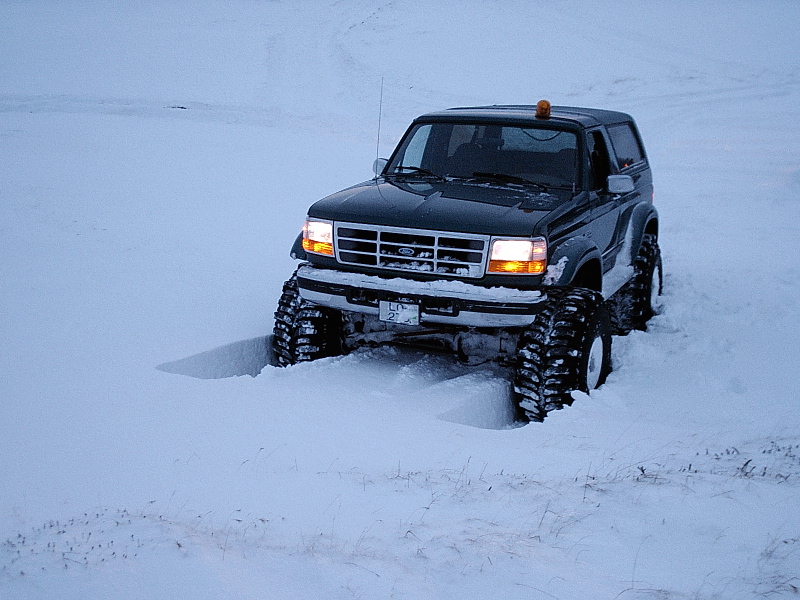 Lifted dark Ford Bronco with big tires driving through deep snow at dusk, front 3/4 view