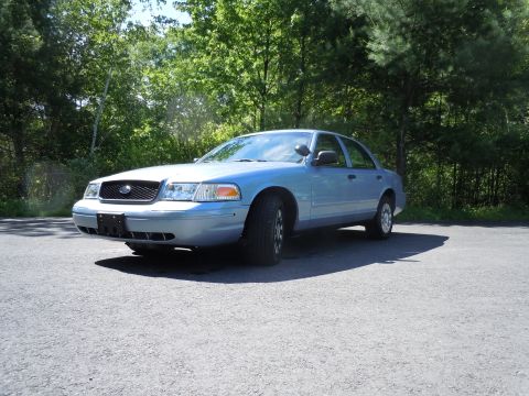 2001 Ford Crown Victoria - A light blue Ford Crown Victoria is parked on an asphalt surface with a backdrop of green trees.