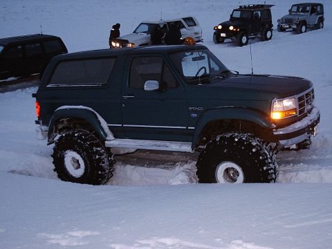 1996 Ford Bronco - A dark green 1996 Ford Bronco navigates deep snow, with other vehicles visible in the background.