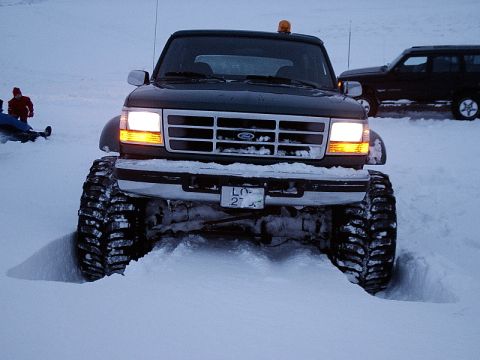 1996 Ford Bronco - 1996 Ford Bronco equipped with oversized tires navigates a snowy landscape, with another SUV visible in the background.