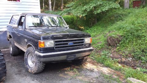 1991 Ford Bronco - A black Ford Bronco with large tires sits parked on a dirt path near a wooded area.