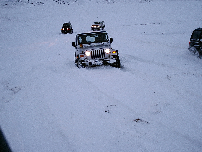 Silver Jeep Wrangler driving through deep snow at dusk with other SUVs nearby