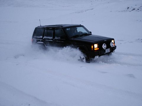 1996 Ford Bronco - A black Jeep Cherokee XJ navigates a snowy terrain, its tires churning up a cloud of snow.