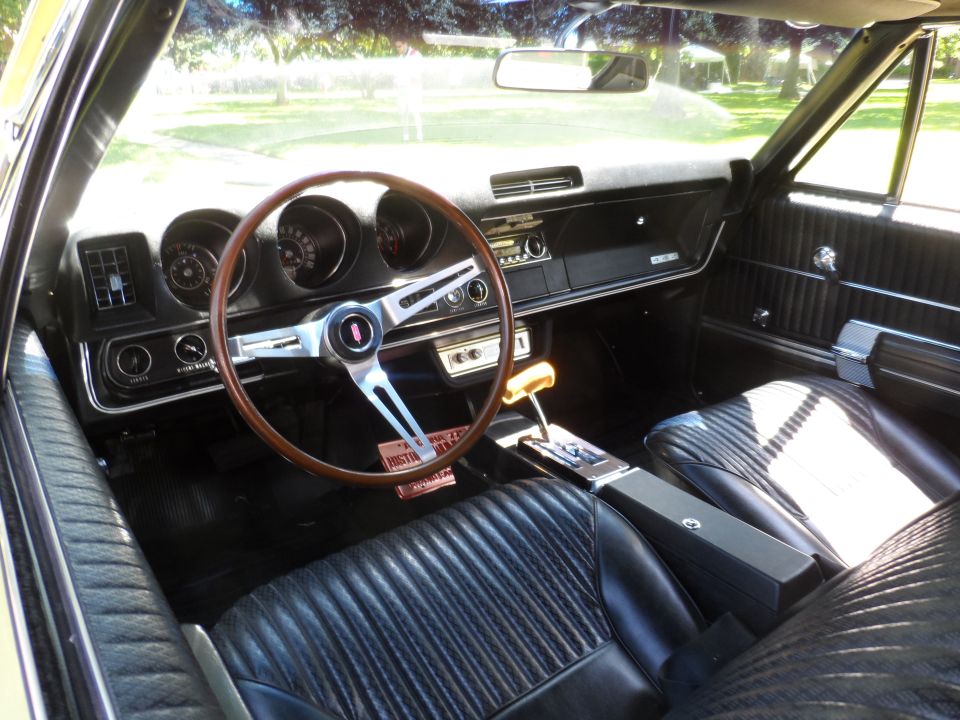 Car interior with black vinyl seats, wood-rimmed steering wheel, and dashboard gauges. A floor shifter is visible