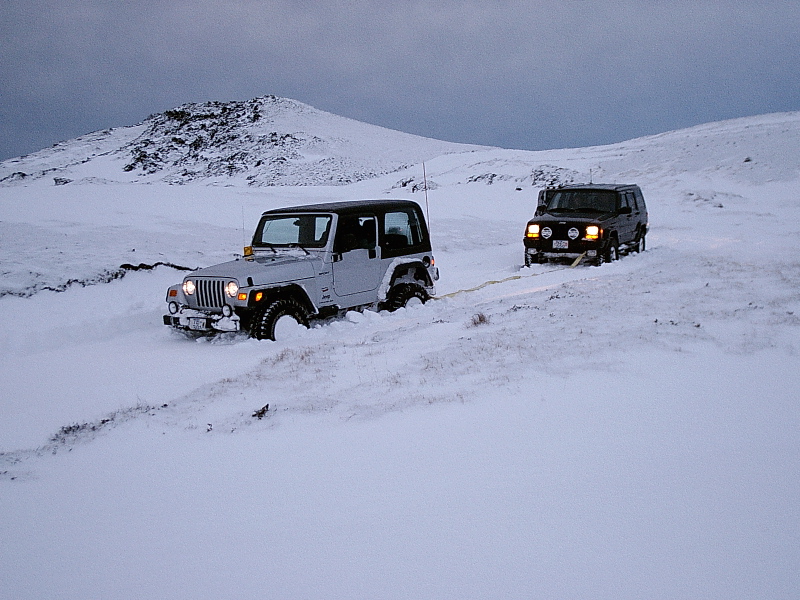 Silver Jeep Wrangler and dark SUV linked by tow strap in deep snow on a hill