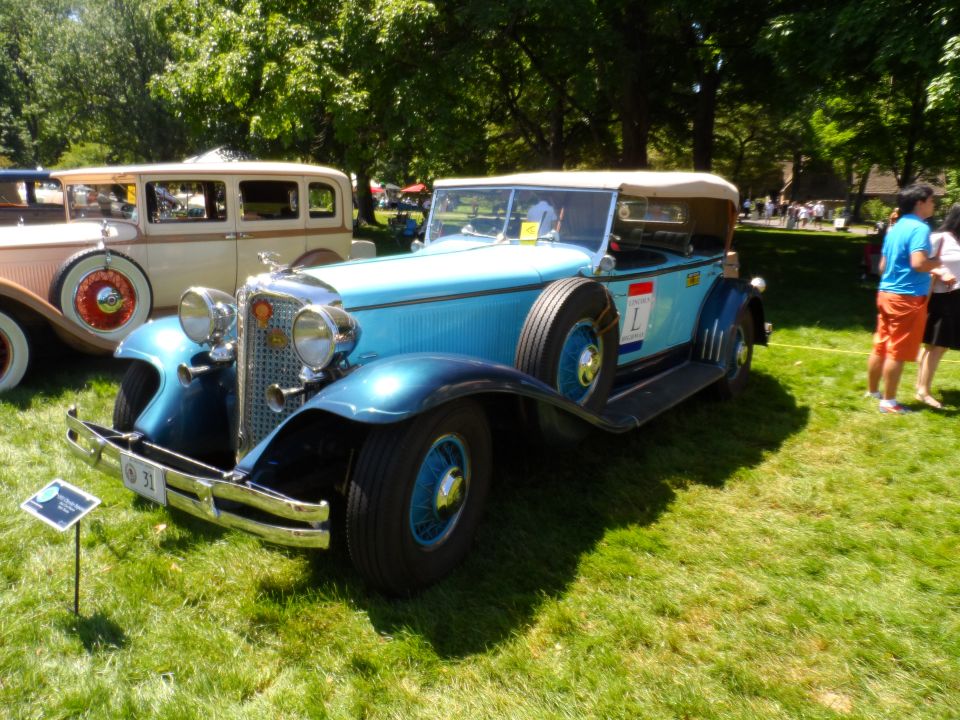 Blue convertible touring car with spare tire on the side, parked on grass next to another vintage car