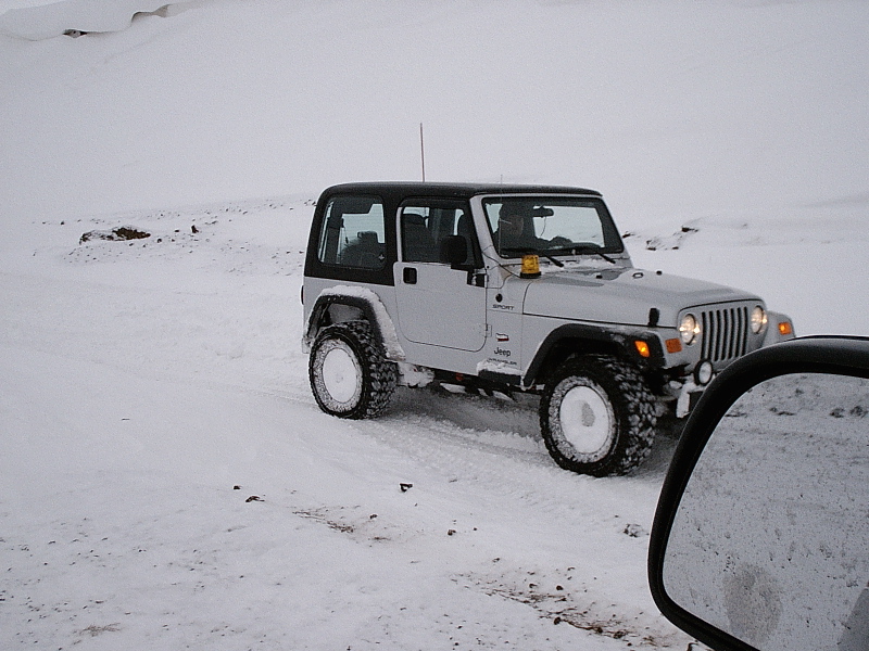Silver Jeep Wrangler with black top driving in deep snow, seen past a car's side mirror