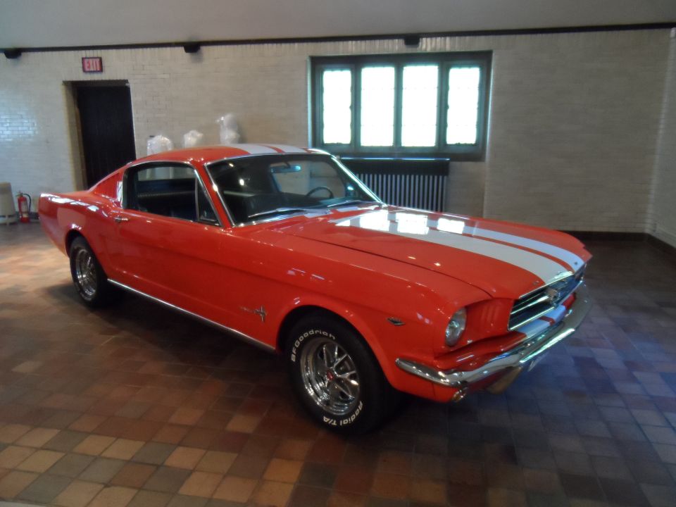 Red 1965 Ford Mustang coupe with white racing stripes, chrome trim, and BFGoodrich tires, viewed from a 3/4 front angle