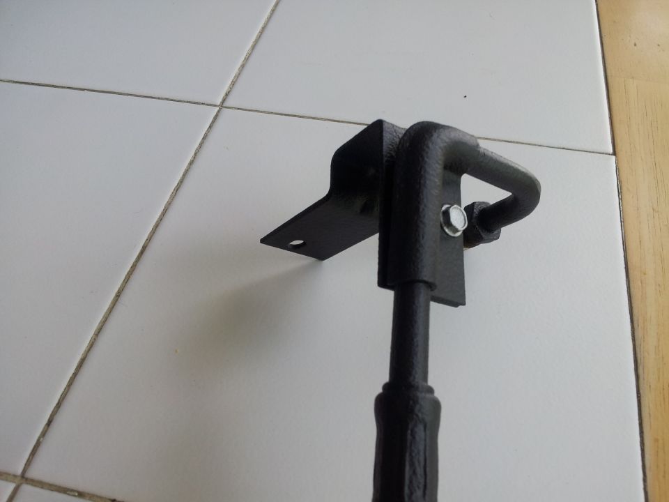 Black metal car part with a hook and mounting bracket, resting on a tiled floor