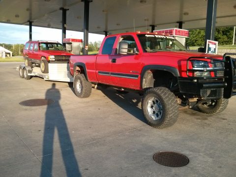 1986 Ford Bronco - Red pickup truck towing a red Jeep Cherokee on a trailer at a gas station.