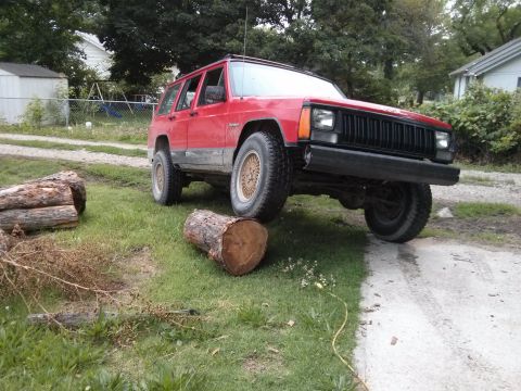 1986 Ford Bronco - A red Jeep Cherokee is parked on a dirt path with logs nearby.