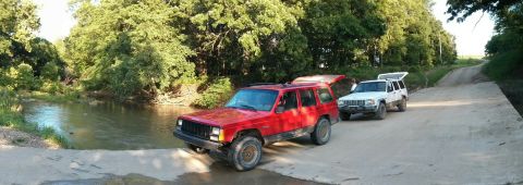 1986 Ford Bronco - Two Jeep Cherokee XJs are parked on a dirt road next to a creek with trees in the background.