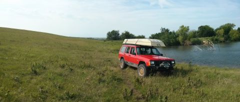 1986 Ford Bronco - Red Jeep Cherokee with a boat on its roof rack parked by a lake on a sunny day.