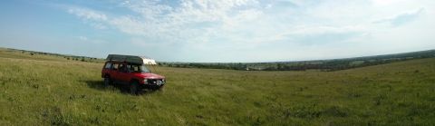 1986 Ford Bronco - A red SUV with a canoe on its roof is parked in a vast, grassy field under a partly cloudy sky.