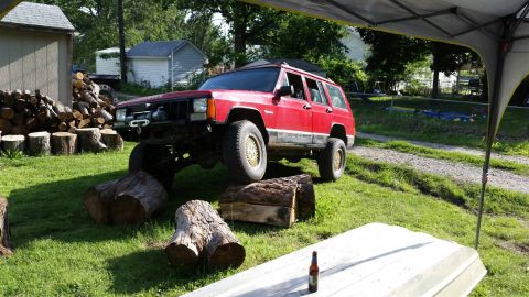 1986 Ford Bronco - Red Jeep Cherokee XJ with a winch and lifted suspension is positioned on logs in a grassy yard.