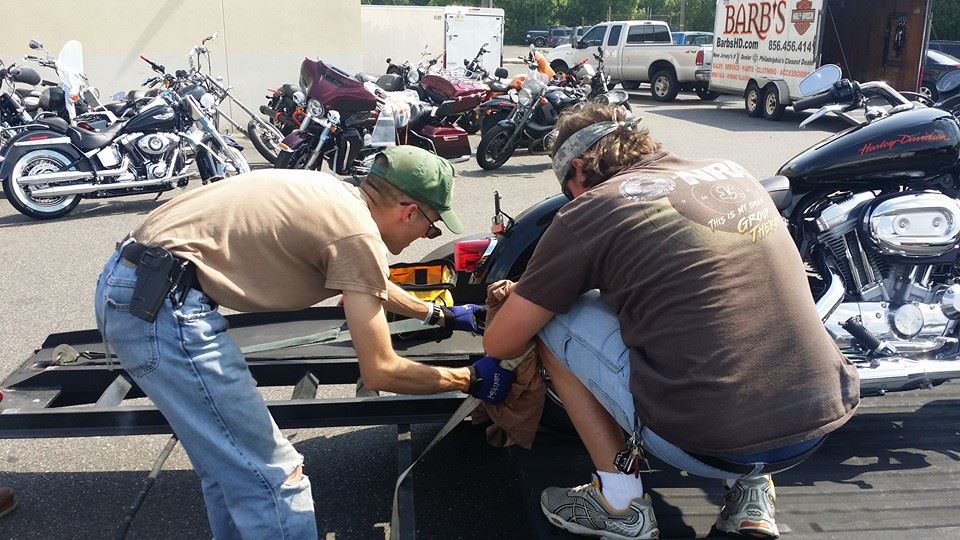 Two men strap a motorcycle to a trailer, with several other motorcycles parked nearby