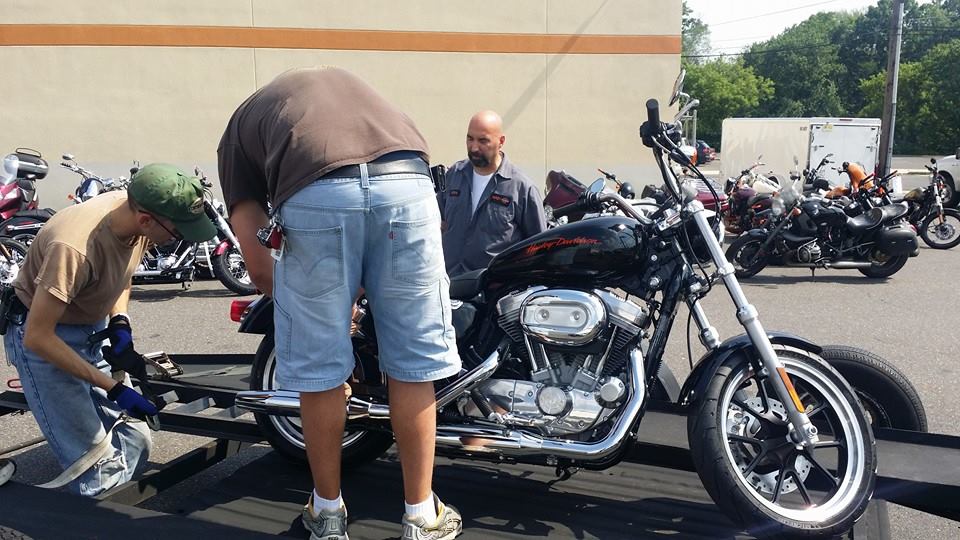 Two men loading a black Harley-Davidson motorcycle onto a trailer, with other motorcycles parked in the background