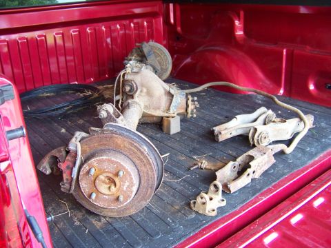 1999 Mercury Grand Marquis - Various rusty axle and suspension parts are loaded into the bed of a red pickup truck.