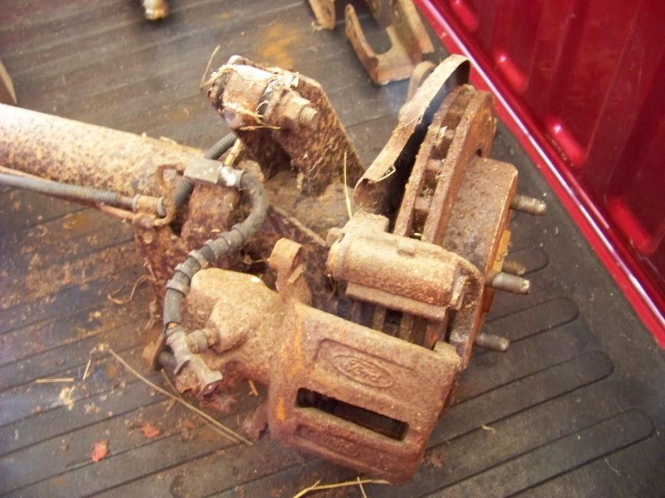 Rusty Ford brake caliper and rotor assembly with brake line attached, resting on a dark surface