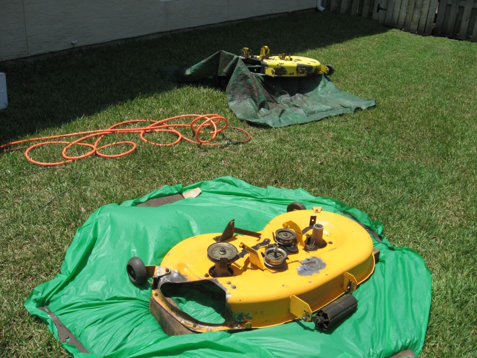 Two yellow lawn mower decks sit on green tarps on a lawn, with an orange extension cord coiled nearby