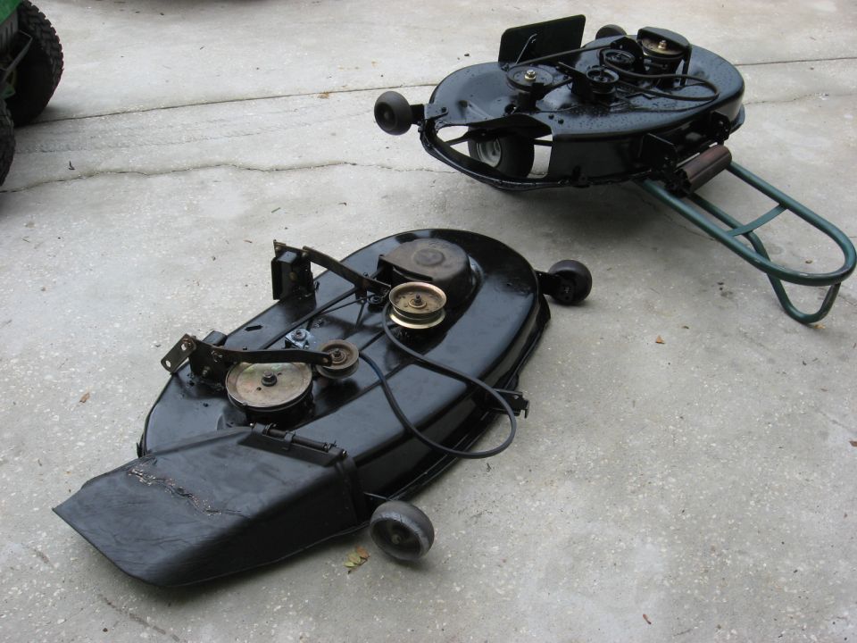 Two black mower decks with pulleys and belts on concrete