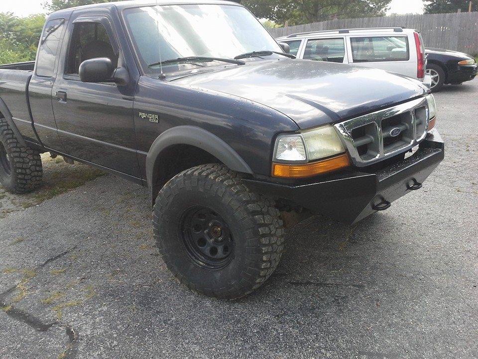 Dark blue Ford Ranger pickup with a custom black front bumper and large tires. A silver SUV and a dark sedan are parked behind it