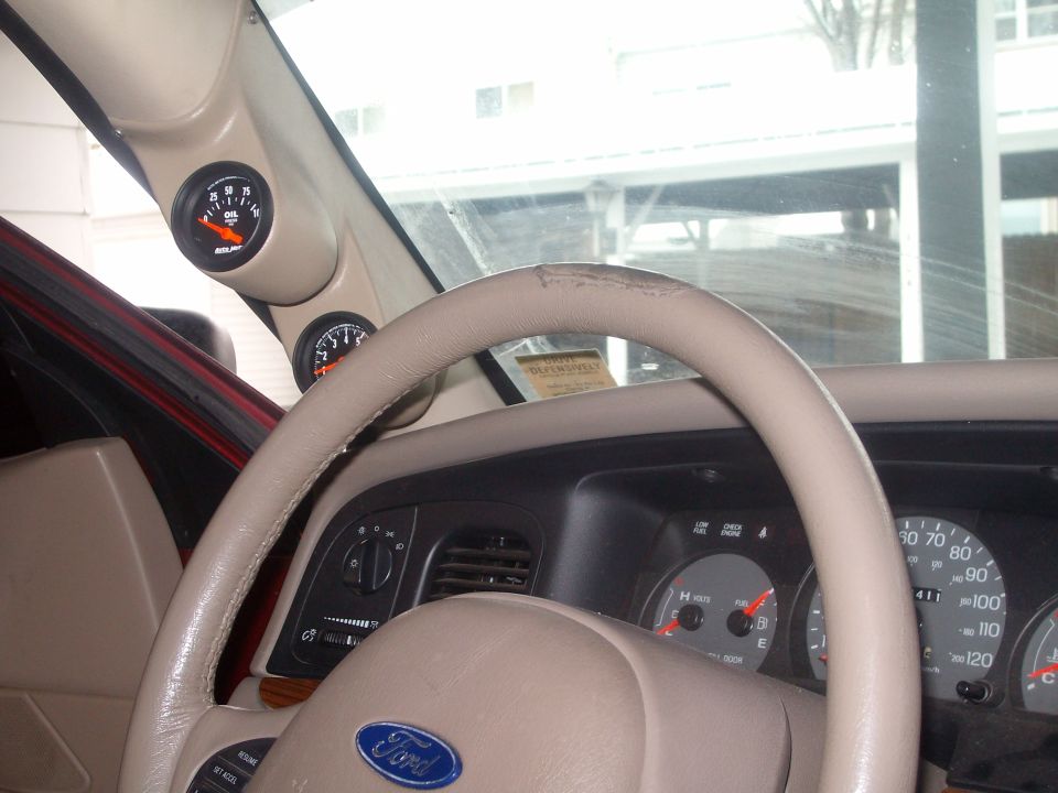 Car interior showing steering wheel, dashboard gauges, and two aftermarket gauges mounted on the A-pillar