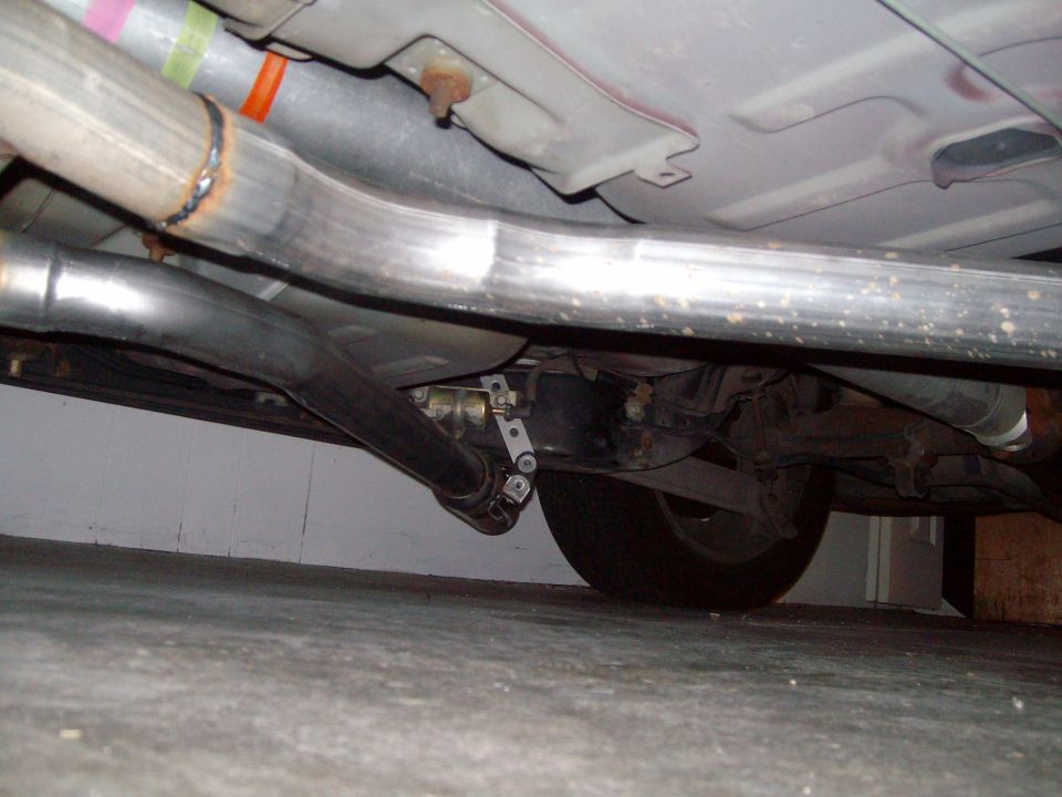 Underside view of a car's exhaust pipes and rear axle. There's a 'Supermotors.net' logo in the corner