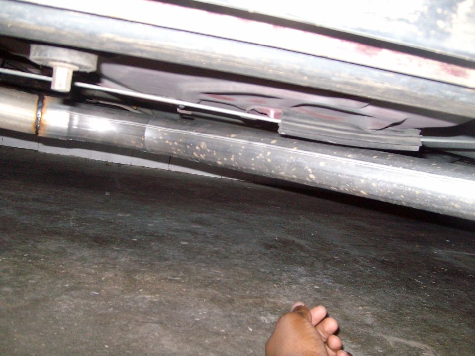 Close-up of a vehicle's exhaust pipe and undercarriage. A hand is visible at the bottom right