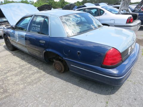 1991 Ford LTD Crown Victoria - A two-tone blue and light blue police car sits in a junkyard with missing wheels and open hoods on other vehicles in the background.