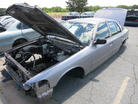 1991 Ford LTD Crown Victoria - A silver 1991 Ford Crown Victoria LTD sits in a lot with its hood and trunk open, showing its dismantled front end and missing wheels.