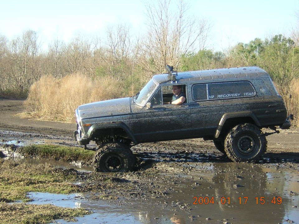 Mud-covered lifted two-door Jeep Cherokee driving through a rutted mud pit, side view